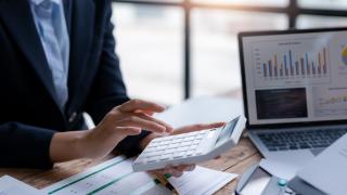 Ein Mann im Anzug sitzt am Schreibtisch und tippt Zahlen in einen Taschenrechner.