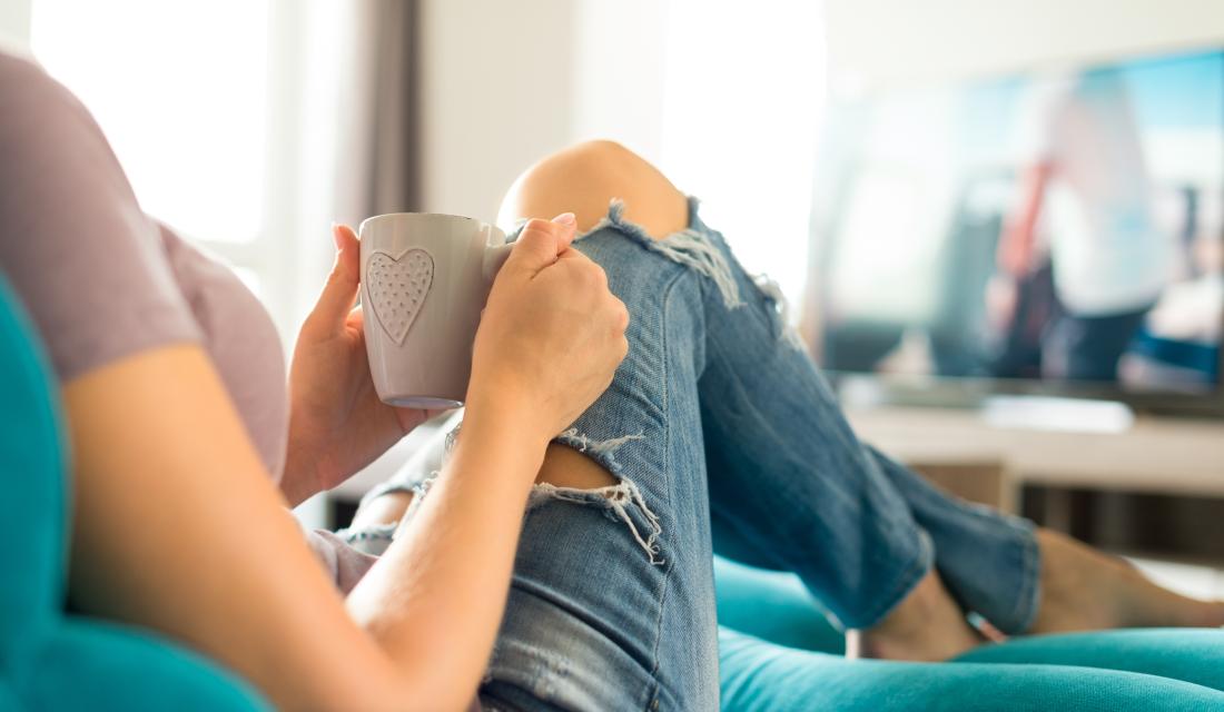 Eine junge Frau sitzt auf dem Sofa, hält eine Tasse in den Händen und sieht fern.